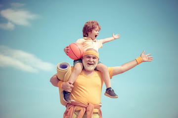 Senior man and cute little boy exercising on blue sky background - isolated. Father and son sporting - family time together. Doing sports is free.
