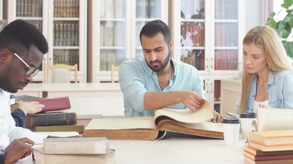 Handsome Arabian young bearded man surrounded by multiracial students holding in hands big ancient book in library.