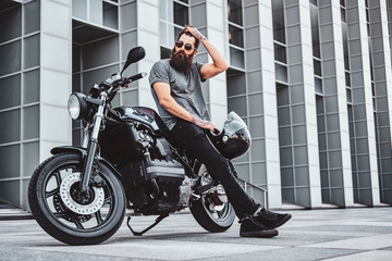 Fototapeta na wymiar Pensive handsome bearded biker is relaxing with his bike near interesting building.