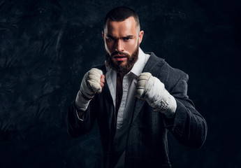 Angry bearded businessman in suit and protective kickboxing gloves is posing for photographer at dark photo studio.
