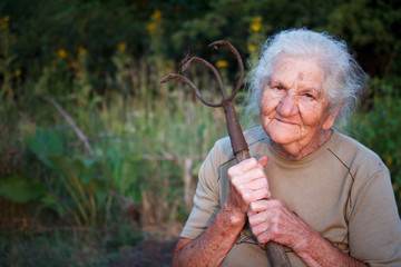 Close-up portrait of an old woman with gray hair holding a rusty pitchfork or chopper in her hands,...