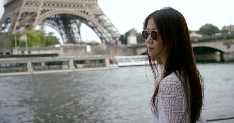 portrait of a dark-haired Asian middle-aged woman close-up, which is located in Paris on the background of the Eiffel tower and the river Seine. She has dark glasses on her face.