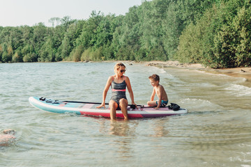 Fototapeta na wymiar Caucasian woman parent sitting on paddle sup surfboard in water with kid child son boy. Modern outdoor family activity. Individual summer aquatic recreation sport hobby. Healthy lifestyle.