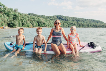 Obraz premium Caucasian woman parent sitting on paddle sup surfboard in water with kids children. Modern outdoor family activity. Individual summer aquatic recreation sport hobby. Healthy lifestyle.