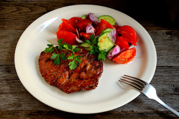 Pork steak, salad tomatoes, cucumbers greens seasonings on a white plate.