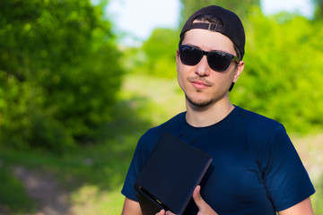 Young guy in glasses and a cap dressed back to front with a laptop on a blurred background of nature