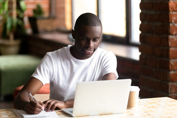 Smiling black male studying on laptop making notes in notebook