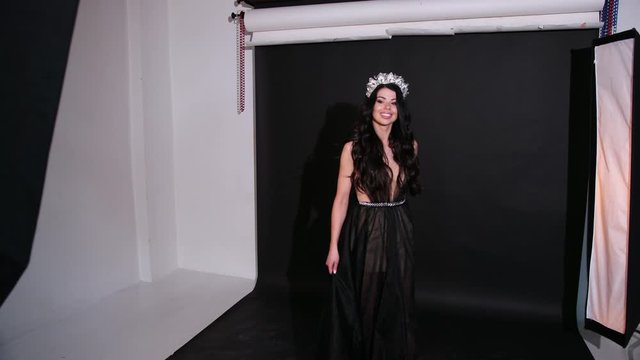 girl model, brunette, in a dark transparent long dress with a deep neckline and naked back, beautiful hair and bright makeup, with a crown on his head, posing in the Studio on a black background,