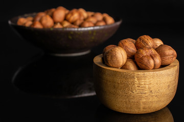 Lot of whole ripe brown hazelnut in dark ceramic bowl in tiny wooden bowl isolated on black glass