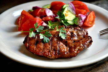 Pork steak, salad tomatoes, cucumbers greens seasonings on a white plate.