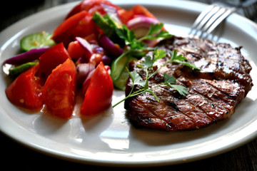Pork steak, salad tomatoes, cucumbers greens seasonings on a white plate.