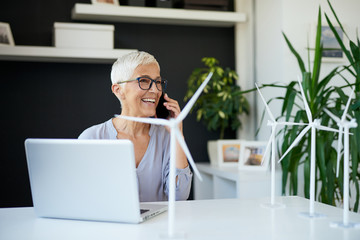 Gorgeous Caucasian senior woman talking on the phone while sitting in office. On desk are are...
