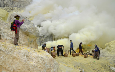 Miners extracting sulfur inside the crater of Kawah Ijen volcano, facing excruciating heat and toxic fumes while a tourist is shooting them.