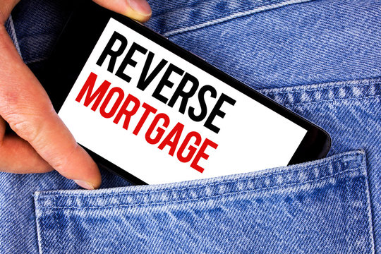 Text sign showing Reverse Mortgage. Conceptual photo Elderly homeowner retirement option regular payment benefit written Mobile phone holding by man the Blue Jeans background.