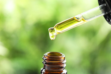 Dripping essential oil from pipette into glass bottle against blurred green background, closeup