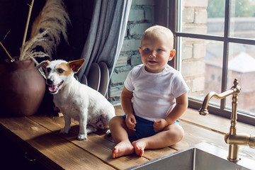 child in a white t-shirt sitting in the kitchen with a dog at the sink