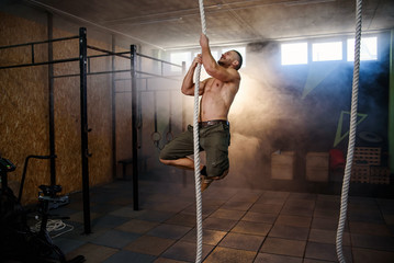 Strong young crossfitter climbing on the rope during training.