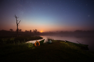 Fototapeta premium Fishermen boats docked at a still lake with sunset in the background. Glowing milky way over the landscape. Tropical summer nights with million stars above and colorful sky. 