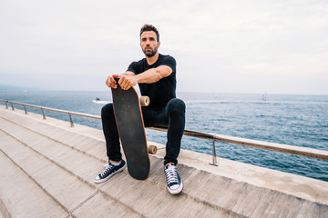 Skateboarder sits with skateboard on the modern city sea terrace. © Igor Kardasov