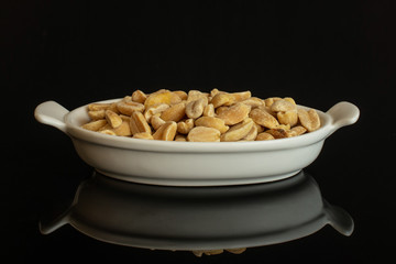 Lot of halves of natural yellow peanut in white oval ceramic bowl isolated on black glass