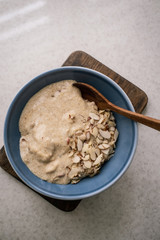 Flaxseed porridge with almonds and coconut milk in a blue bowl, wooden spoon on white marble table