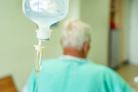 Asian Patient Older Man Lying At Hospital Bed With Saline Drip Showing Victory Sign. Drop Of Saline Solution To Help Patient, Iv Fluid Use For Intravenous Volume. Selective Focus