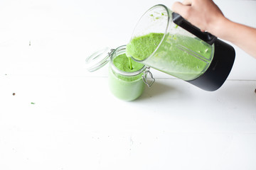Female hand pouring a healthy green smoothie from the blender in a glass jar on a white wooden background