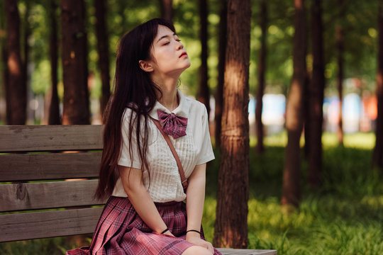 Portrait of beautiful Chinese girl in white shirt and pink skirt sitting on bench in sunny summer forest, breathing the fresh air with eyes closed. Beauty, lifestyle and summer Concept.
