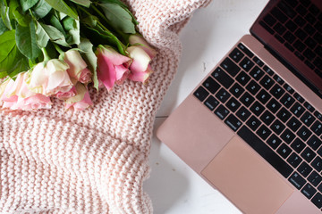 Womens workspace with laptop, bouquet of pink roses, a notebook, a pen and a cosy blanket in a flat lay top view