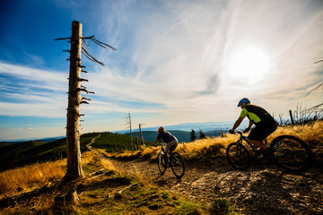 Cycling woman and men riding on bikes at sunset mountains forest landscape. Couple cycling MTB enduro flow trail track. Outdoor sport activity.