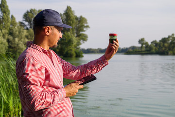 ecologist on river bank examines container with green algae and holds tablet