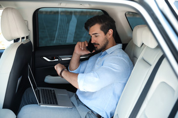 Attractive young man working with laptop and talking on phone in luxury car