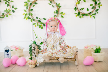 Fototapeta na wymiar Cute little girl in a dress with a flower print and a bunny hat in the Easter decorations in the studio. Little girl with easter eggs and flowers in a spring studio