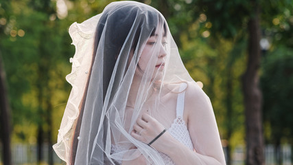 Portrait of beautiful Chinese girl in white bride dress looking away with veil on head in windy dusk with blur forest background, Beauty, lifestyle and summer Concept.