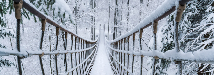 Im verschneiten Winterwald hängt eine Seilbrücke. Verschneite Tannenbäume säumen den Pfad....