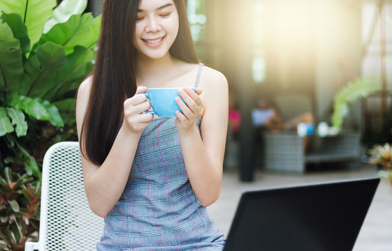 Portrait of young asian woman holding blue cup of coffee to drink when she working with laptop in outdoor cafe - Powered by Adobe