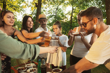 Group of happy friends eating and drinking beers at barbecue dinner on sunset time. Having meal together outdoor in a forest glade. Celebrating and relaxing. Summer lifestyle, food, friendship concept
