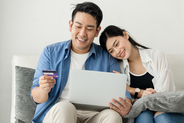 Fototapeta premium Happy Asian couple shopping online with holding credit card and laptop and sitting at home together, lifestyle