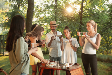 Group of happy friends eating and drinking beers at barbecue dinner on sunset time. Having meal together outdoor in a forest glade. Celebrating and relaxing. Summer lifestyle, food, friendship concept