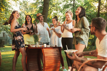 Group of happy friends eating and drinking beers at barbecue dinner on sunset time. Having meal together outdoor in a forest glade. Celebrating and relaxing. Summer lifestyle, food, friendship concept