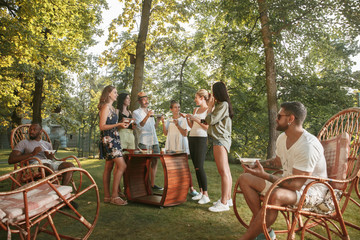 Group of happy friends eating and drinking beers at barbecue dinner on sunset time. Having meal together outdoor in a forest glade. Celebrating and relaxing. Summer lifestyle, food, friendship concept