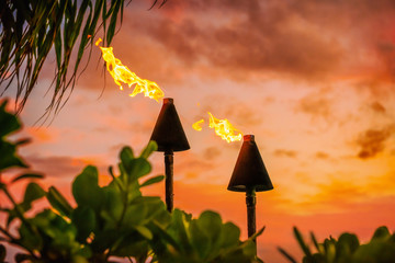 Hawaii luau party Maui fire tiki torches with open flames burning at sunset sky clouds at night. Hawaiian cultural travel vacation background. - obrazy, fototapety, plakaty