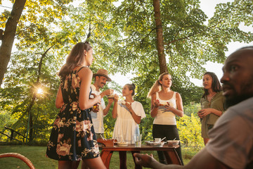 Fototapeta premium Group of happy friends eating and drinking beers at barbecue dinner on sunset time. Having meal together outdoor in a forest glade. Celebrating and relaxing. Summer lifestyle, food, friendship concept