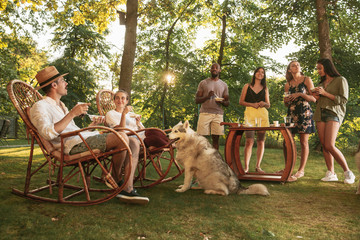 Fototapeta na wymiar Group of happy friends eating and drinking beers at barbecue dinner on sunset time. Having meal together outdoor in a forest glade. Celebrating and relaxing. Summer lifestyle, food, friendship concept