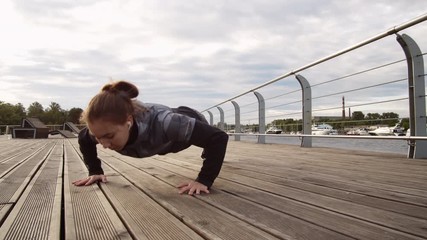Pan shot of young sporty Caucasian brunette doing push-ups on wooden embankment outdoors