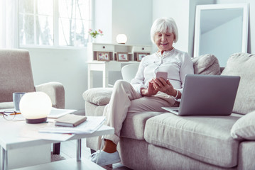 Elderly businesswoman sitting in living room with smartphone and laptop