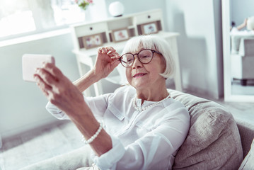 Naklejka na ściany i meble Stylish appealing retired woman holding her glasses while making selfie
