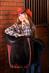 A sexy girl is standing in a stables dressed in a cowboy hat, cowboy boots and jeans. 
