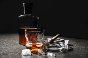 Glass and bottle of cold whiskey with ice and cigar on dark table