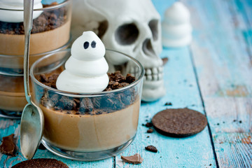 Halloween dessert for kids - delicious chocolate mousse with chocolate cookie crumbs and meringue ghost in glass on table with festive halloween decorations